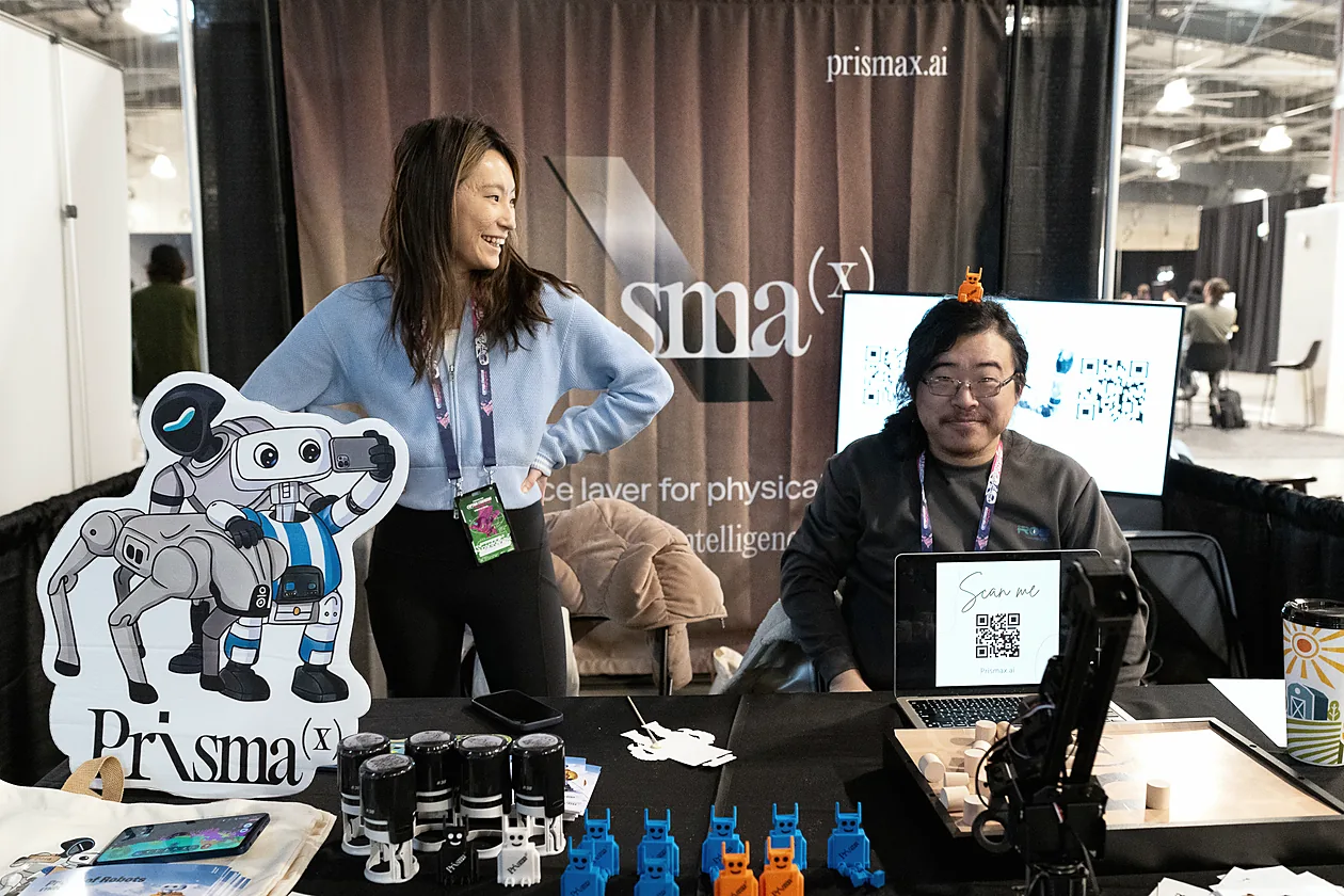 Two PrismaX team members at the PrismaX booth at ETHDenver, with a robot arm demo and robot figurines on the table