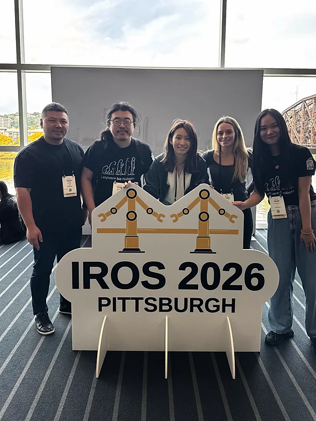 Five PrismaX team members in PrismaX shirts behind the IROS 2026 Pittsburgh sign
