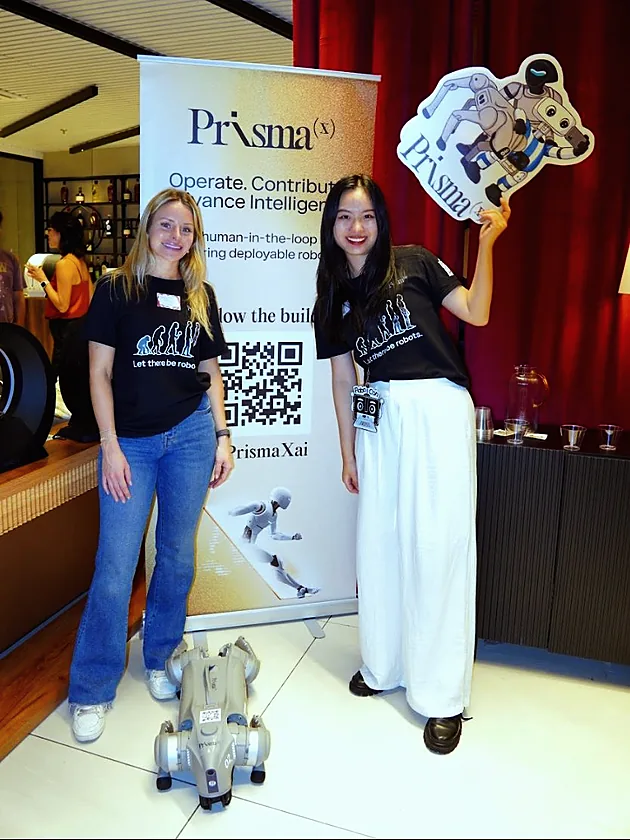 Two PrismaX team members by a PrismaX banner at a16z Tech Week, one holding up a PrismaX robot sign